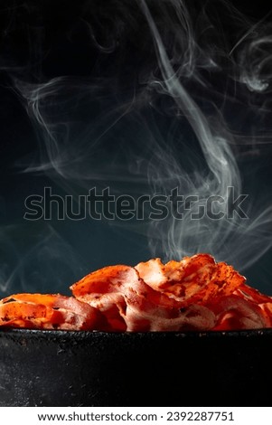 Fried steaming bacon slices in an old black pan on a black background. Copy space.