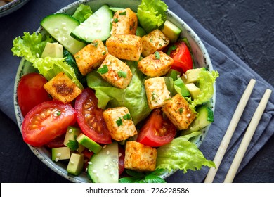 Fried Tofu Salad With Cucumbers, Tomatoes, Avocado And Sesame Seeds. Homemade Asian Vegetable And Tofu Salad In Ceramic Bowl On Black Stone Background. Healthy Asian Diet Vegan Vegetarian Salad Food.