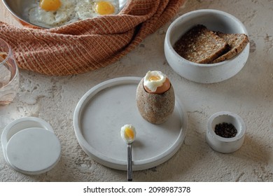 Fried two eggs in copper pan, hard boiled egg in stoneware egg cup, salt flakes, pepper, whole grain rye bread in concrete bowl on rough textured clay background. Healthy breakfast concept. - Powered by Shutterstock