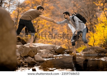 Friend giving a helping hand while crossing the stream barefooted walking by the creek with his shoes in hand adventure travel hiking trail forest. 