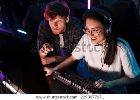 Friend of young emotional asian girl watching her playing online video game and assisting her in modern cybersport club