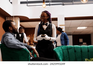 Friendly African American female waitress taking order serving hotel guest in lobby. Young black man traveler ordering coffee while waiting for check-in. Customer service in hospitality industry - Powered by Shutterstock