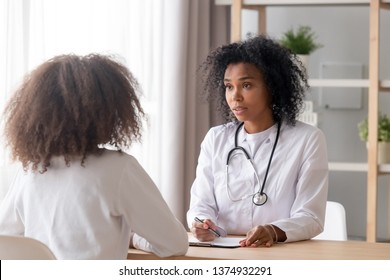 Friendly African American Pediatrician Doctor In White Uniform With Stethoscope Talking To Teen Girl During Visit, Listening To Complaint At Consultation, Fill Patient Form At Medical Checkup