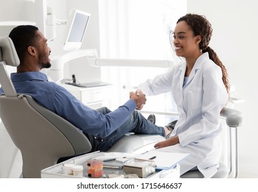 Friendly african woman dentist shaking male patient hand sitting in dentist chair, empty space - Powered by Shutterstock