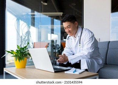 Friendly Asian Doctor Uses Laptop To Remotely Communicate With Patient, Man In Clinic Consults Online, Looks At Monitor Screen And Webcam