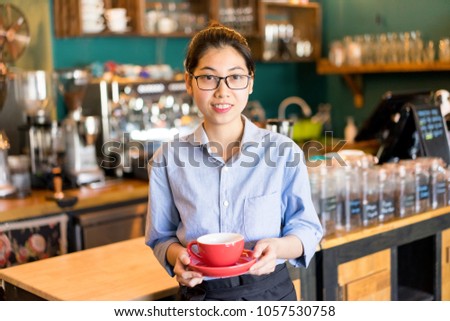Friendly Asian female waitress holding coffee cup and looking at camera. Cheerful beautiful young barista making perfect coffee. Cafe staff concept