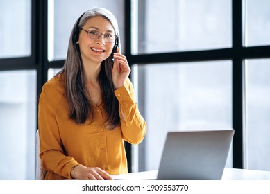 Friendly asian mature gray-haired business woman or tutor, in casual stylish wear, headset and eyeglasses is working online, or conduct distance teaching, using laptop at workplace, looks at the