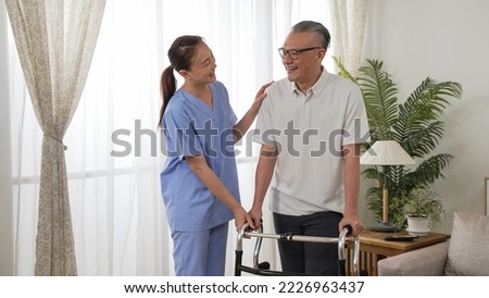friendly asian personal care attendant chatting with mature male patient while he is standing with a walker near the window at home
