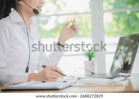 Friendly Asian receptionist smiling while talking through headset in modern office, helping customers and supporting coworkers with professional teamwork.