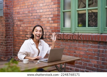 Friendly asian woman sitting with laptop, waving at you, saying hello, hi gesture, greeting you while working with computer.