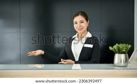 Friendly and attractive millennial Asian female receptionist in a uniform stands at a front desk in modern lobby. Hospitality service concept 商業照片 © 