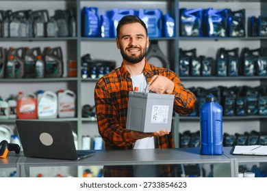 Friendly bearded man working in auto shop, holding a car battery, with shelves of oil and automotive products in background