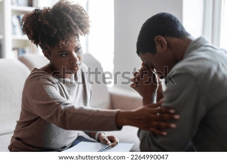 Friendly beautiful black woman therapist consoling upset young man, african american female psychologist touching sad crying patient shoulder, clinic interior. Psychological support concept