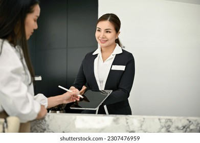 A friendly and beautiful young Asian female receptionist is talking and handing a digital tablet to a customer to sign at the front desk. Hotel check-in, Hospitality service concept