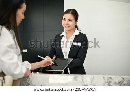 A friendly and beautiful young Asian female receptionist is talking and handing a digital tablet to a customer to sign at the front desk. Hotel check-in, Hospitality service concept