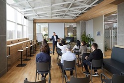 Friendly Business Coach Communicates With Group Of Adult Students At Seminar In Advanced Training. Senior Businessman Answers Questions From Listeners Sitting At Desks In Office With Loft Interior.