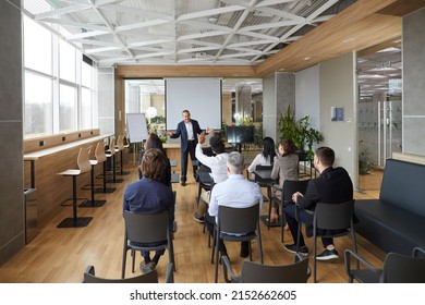 Friendly Business Coach Communicates With Group Of Adult Students At Seminar In Advanced Training. Senior Businessman Answers Questions From Listeners Sitting At Desks In Office With Loft Interior.