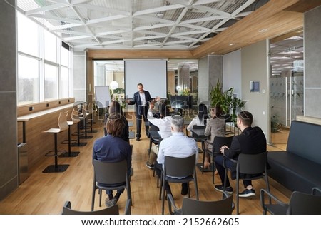 Friendly business coach communicates with group of adult students at seminar in advanced training. Senior businessman answers questions from listeners sitting at desks in office with loft interior.