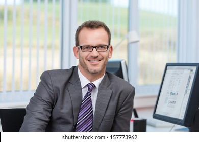 Friendly businessman wearing glasses sitting at his desk in the office looking at the camera with a smile