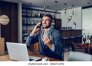 A Friendly Busy Man Is Sitting In The Working Friendly Cafe And Having A Phone Call With Colleagues. There Is A Laptop On A Table. A Businessman Talking On The Phone In A Cafe