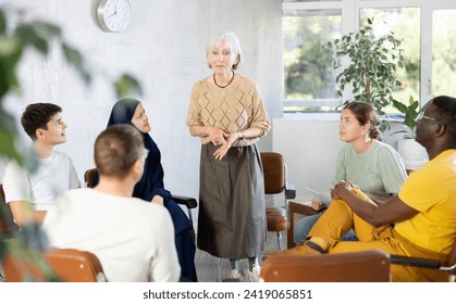 Friendly and calm senior female psychotherapist or facilitator leading group therapy session with women and men of different ages and nationalities sitting  - Powered by Shutterstock