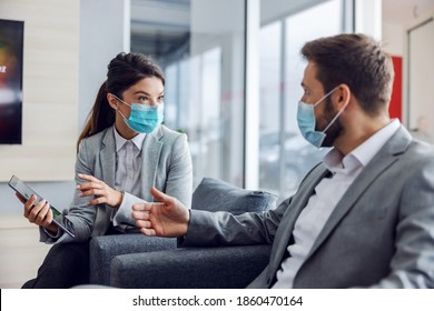 Friendly Car Seller With Face Mask Sitting With Customer In Car Salon, Holding Tablet And Talking About Specifications And Performances Of Car During Corona Virus.