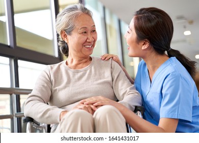 Friendly Caregiver Of Nursing Home Talking To Asian Senior Woman In Hallway