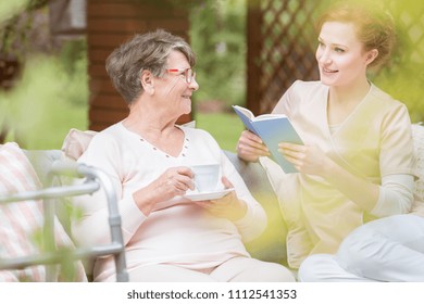 Friendly Caregiver Reading A Book And Senior Woman Drinking Tea In The Garden