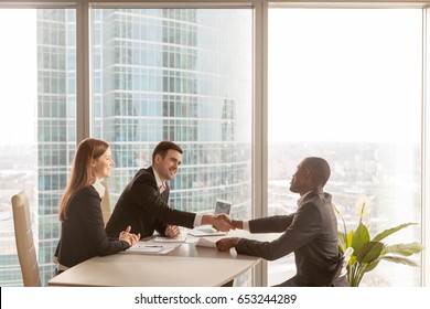 Friendly caucasian employers and confident african-american applicant handshaking during job interview sitting at office desk with big large window urban buildings cityscape at background, side view