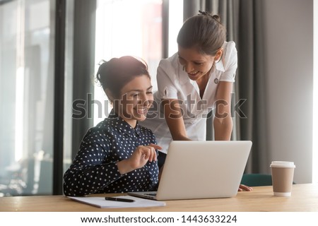 Friendly caucasian female mentor manager supervise indian intern secretary helping coworker with computer work instruct trainee give advice at office desk, apprentice, teamwork and mentoring concept