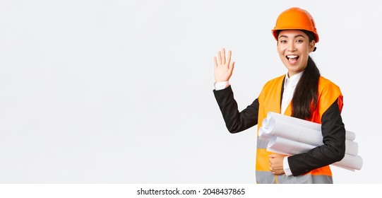 Friendly Cheerful Asian Female Construction Manager, Lead Architect In Safety Helmet And Jacket, Carry Blueprints, Documents Of Building Project, Waving Hand To Say Hello, Greeting Someone