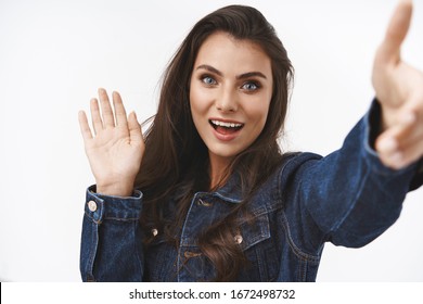 Friendly and cheerful, beautiful brunette fashion blogger girl, saying hi to followers, greeting viewer, wave hand hello gesture, taking selfie, record video using smartphone over white background
