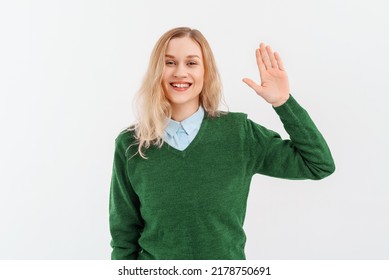 Friendly Cheerful Blonde Girl Smiling, Saying Hi And Waving Hand To Greet Person, Make Hello Gesture, Welcome Someone, Wears Casual Clothes. Indoor Studio Shot On White Background