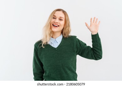 Friendly Cheerful Blonde Girl Smiling, Saying Hi And Waving Hand To Greet Person, Make Hello Gesture, Welcome Someone, Wears Casual Clothes. Indoor Studio Shot On White Background