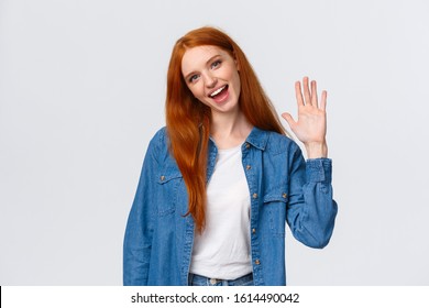 Friendly, cheerful and outgoing pretty redhead girl waving palm, saying hello, greeting team member, tilt head and smiling joyfully, nice meet you, say hi and standing white background