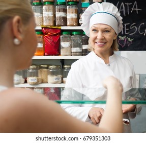 Friendly Cheerful Shop Woman Offering Senior Client Nuts And Smiling In Shop