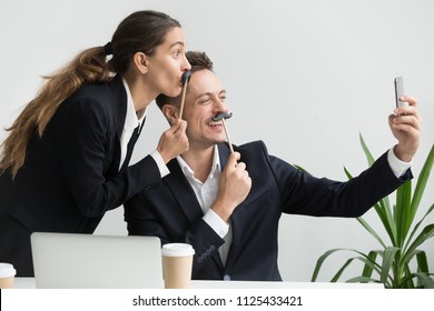 Friendly colleagues in suits having fun grimacing holding fake mustache taking picture on smartphone in office, happy coworkers making funny faces laughing at phone camera, silly selfie concept - Powered by Shutterstock