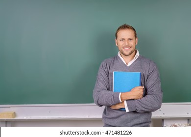 Friendly Confident Post Graduate Student Standing Clutching A Folder Of Class Notes With His Back To A Blank Blackboard With Copy Space