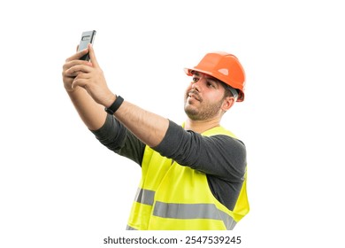 Friendly constructor wearing fluorescent work vest and orange safety hardhat taking selfie using smartphone camera isolated on white studio background