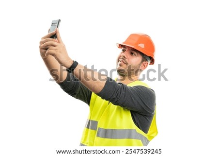 Friendly constructor wearing fluorescent work vest and orange safety hardhat taking selfie using smartphone camera isolated on white studio background