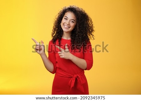 Friendly cute assertive female assistant in elegant red dress with curly hair tilting head flirty and smiling broadly, satisfied as pointing at upper left corner, recommending product over yellow wall