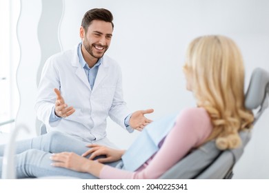 Friendly Dentist Handsome Millennial Man In Workwear Having Conversation With Blonde Woman Patient Sitting At Dental Chair, Smiling And Gesturing, Side View. Doctor And Patient Relationship