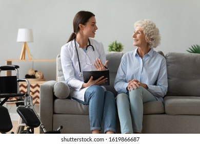 Friendly Doctor Consulting Disabled Woman, Giving Prescription, Holding Clipboard, Discussing Good Medical Checkup Results At Home, Smiling Nurse And Happy Mature Patient Chatting, Sitting On Couch