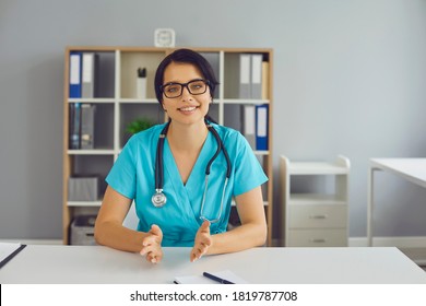 Friendly Doctor At Desk Looking At Camera And Providing Consultation To Patient During Video Call. General Practitioner In Hospital Office Talking To Client Or Making Professional Vlog Or Webinar