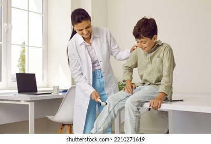 Friendly Doctor Examining Little Child. Professional Neurologist Uses Hammer To Check Knee Reflex Of Happy School Boy Sitting On Medical Couch In Exam Room. Children, Health Checkup, Neurology Concept