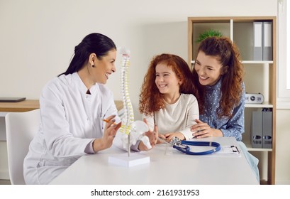 Friendly doctor shows little girl anatomical model of spine standing on table in doctor's office. Joyful mother and her cute redhead little daughter at reception of modern children's clinic. - Powered by Shutterstock