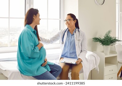 Friendly doctor supports pregnant woman patient at clinic. Smiling nurse or obstetrician with clipboard puts her hand on shoulder of young pregnant lady. Pregnancy, health care concept - Powered by Shutterstock