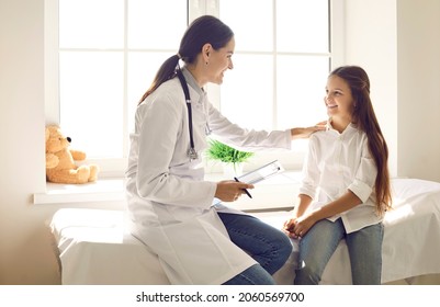 Friendly Doctor Talking To Child And Touching Her Shoulder To Support Her At Medical Checkup. Happy Teen Girl Sitting On Examination Couch In Exam Room And Listening To Female Pediatrician's Advice