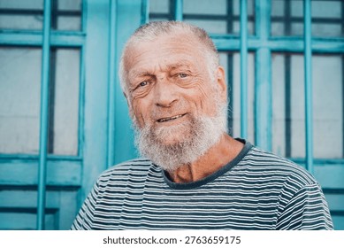 Friendly elderly man with a white beard smiles warmly in a close-up portrait against a turquoise windowed background, conveying warmth, wisdom, resilience and relaxed seaside or coastal vibe.
