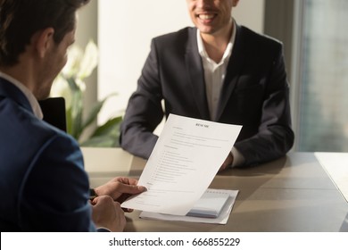 Friendly employer conducting job interview, reviewing good resume of prepared skilled smiling applicant waiting for result at background, recruiter considering cv, focus on document, close up view 